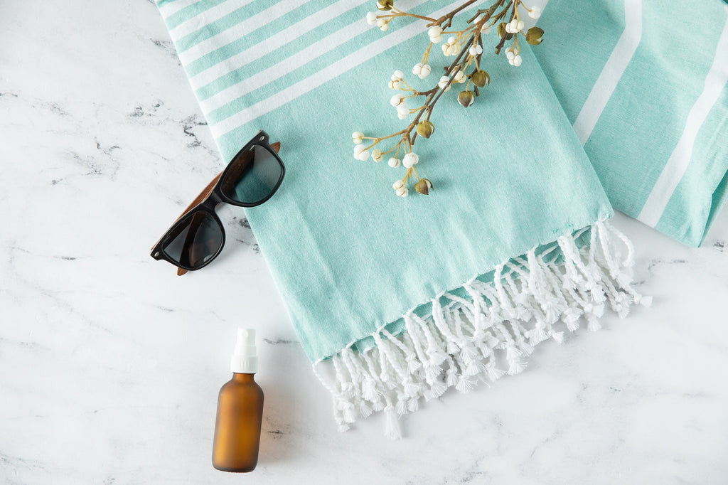 Turquoise towel with white stripes and fringes, sunglasses, and a bottle on a light surface.
