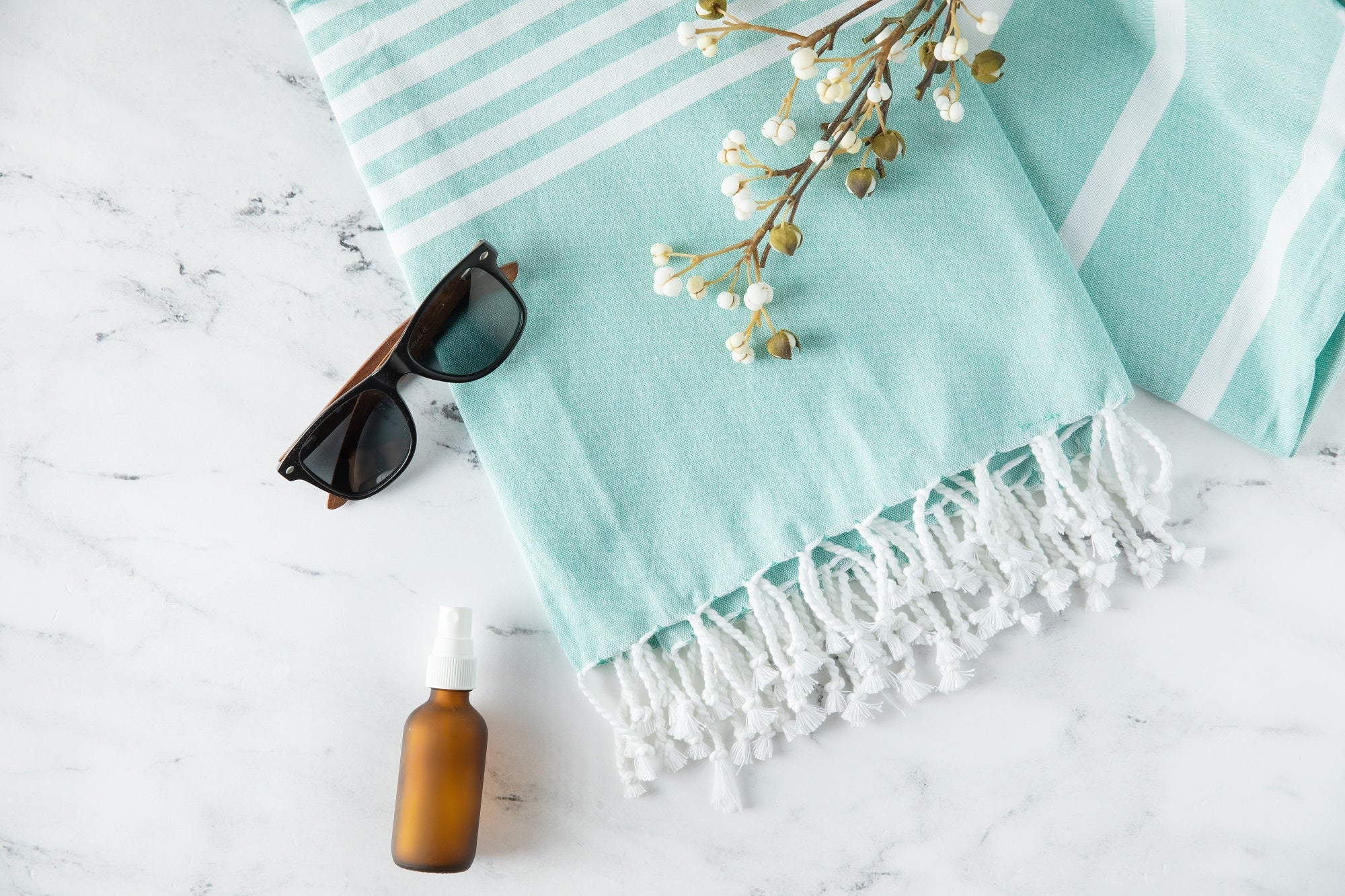 Turquoise towel with white stripes and fringes, sunglasses, and a bottle on a light surface.