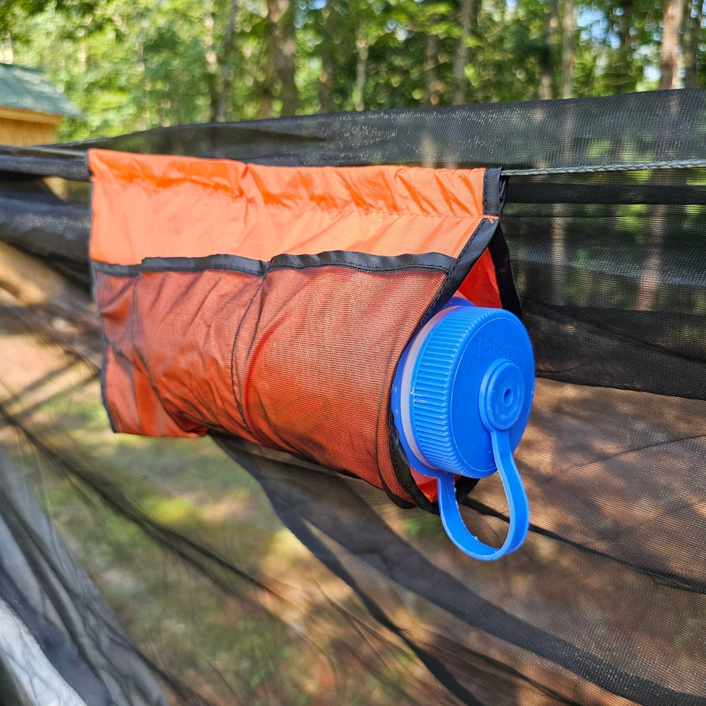 Orange mesh bag with a blue lid hanging on a black mesh screen with a forest background.